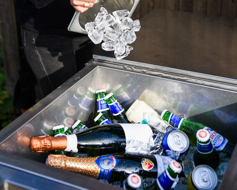 Person adding ice to a cooler filled with various bottles outdoors.